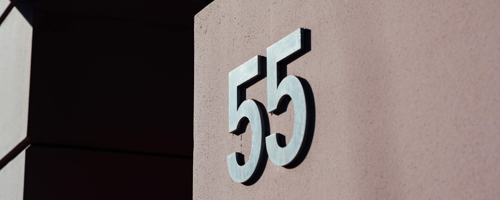 Close-up of a modern building wall with metal number 55 casting a shadow.